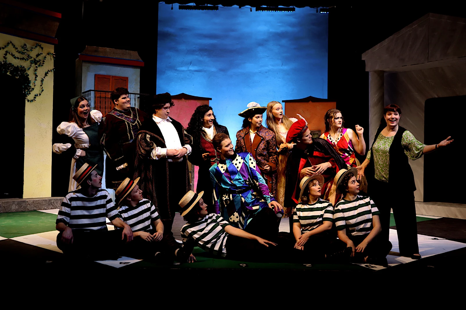 An ensemble of costumed performers gathers on a theater stage in front of a stylized town backdrop during a group scene.