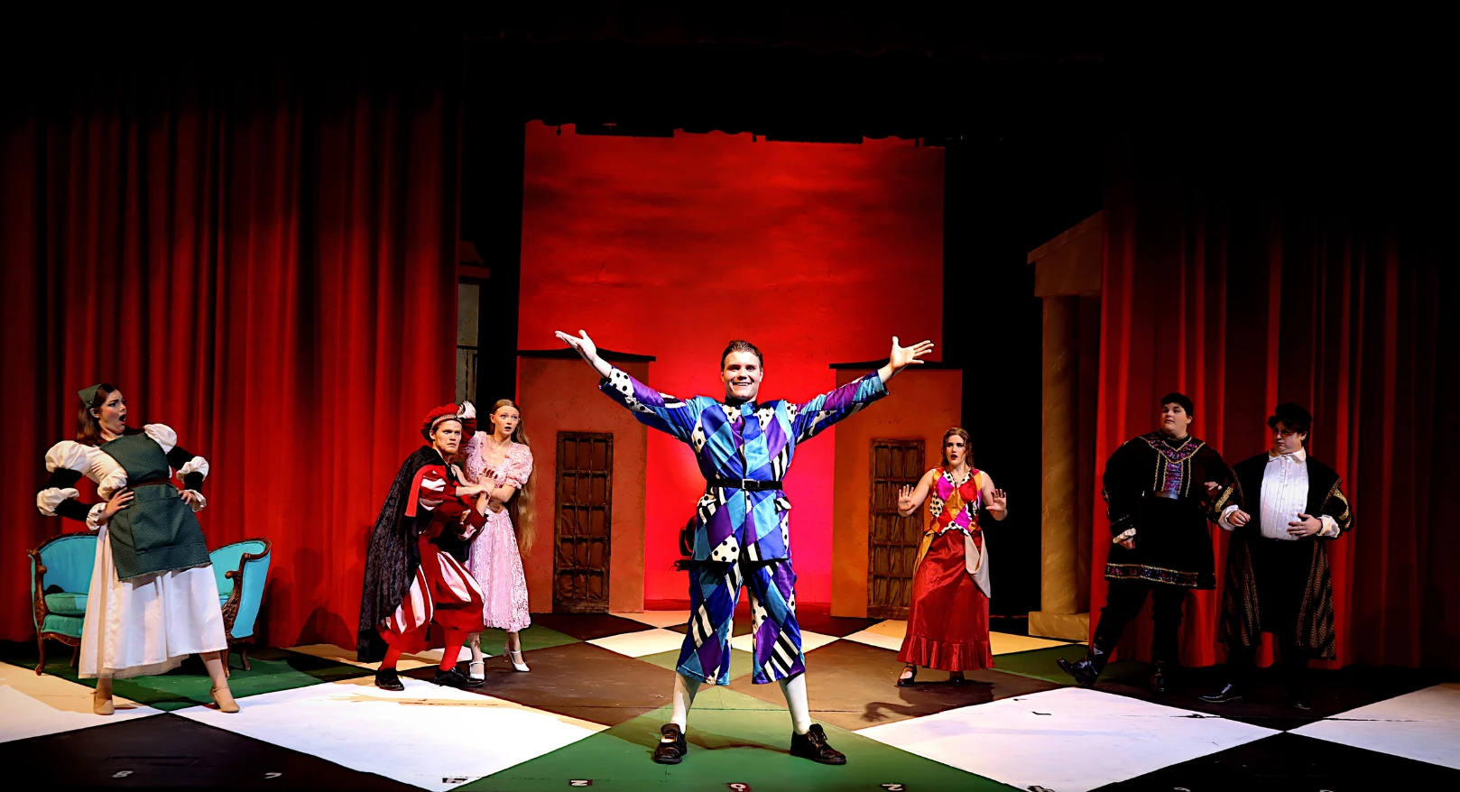 A performer in a blue patterned costume stands center stage with arms outstretched while other cast members surround the scene beneath red stage lighting.