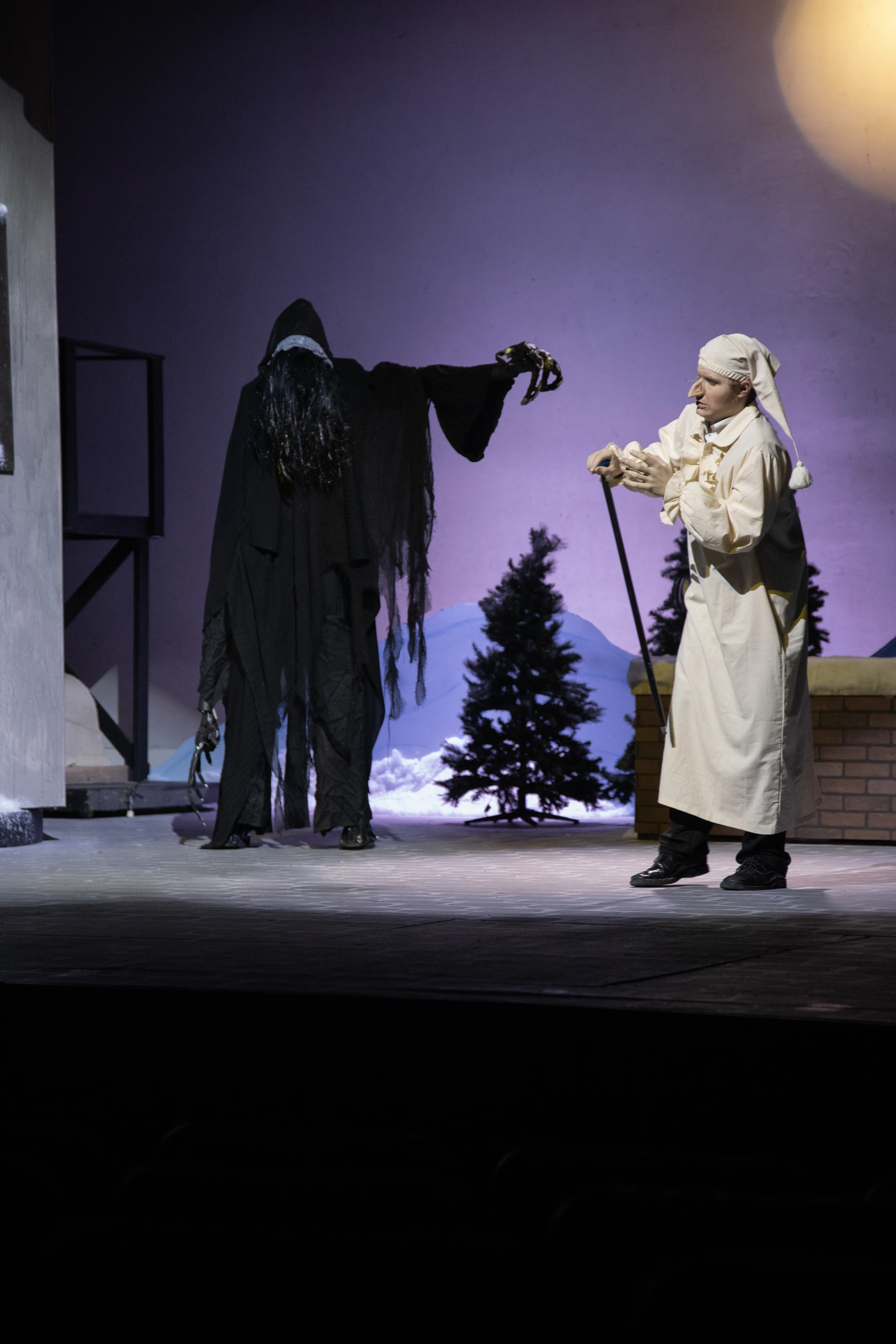 A performer in a white nightgown confronts a dark, hooded figure on a winter-themed stage set during a theatrical scene.