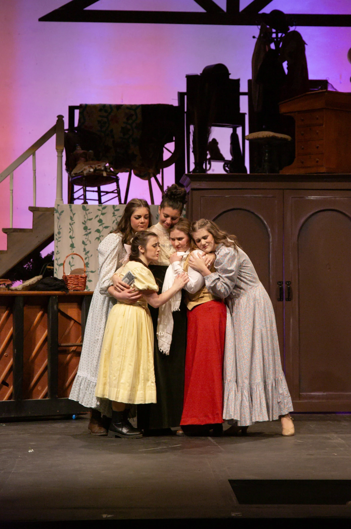 Five performers in period-style clothing stand closely together on a stage set resembling a family home.