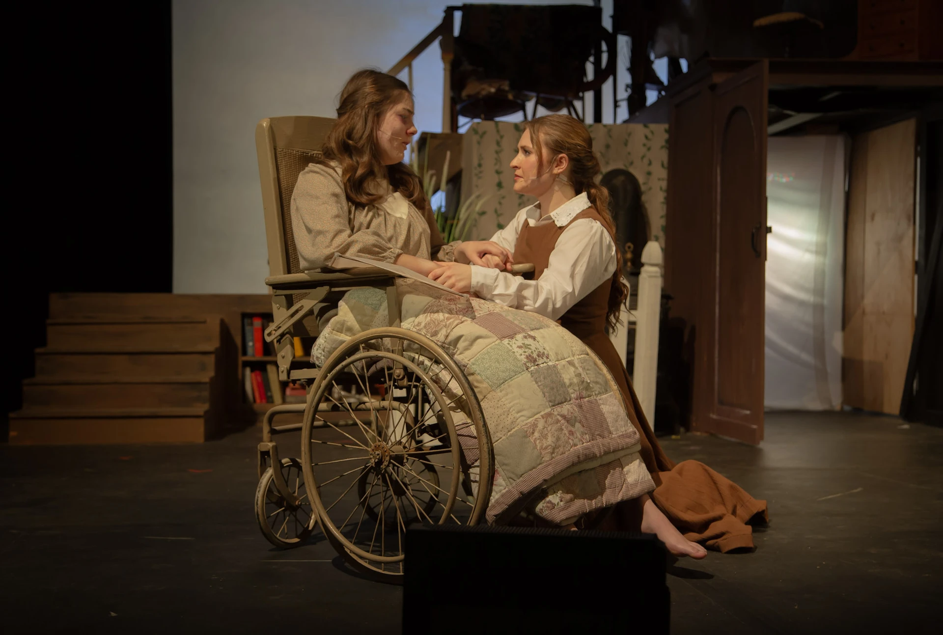 One performer seated in a wheelchair faces another performer kneeling beside them on a theater stage with a domestic interior set.