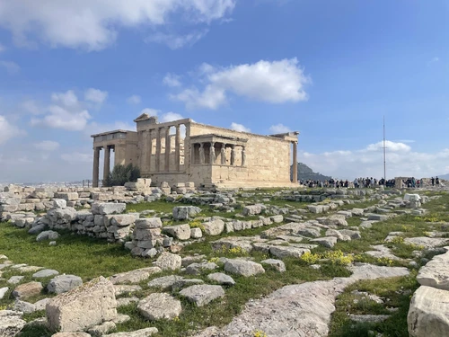 Ancient stone ruins of the Erechtheion temple on the Acropolis in Athens, Greece, with scattered stones in the foreground and a partly cloudy blue sky above.