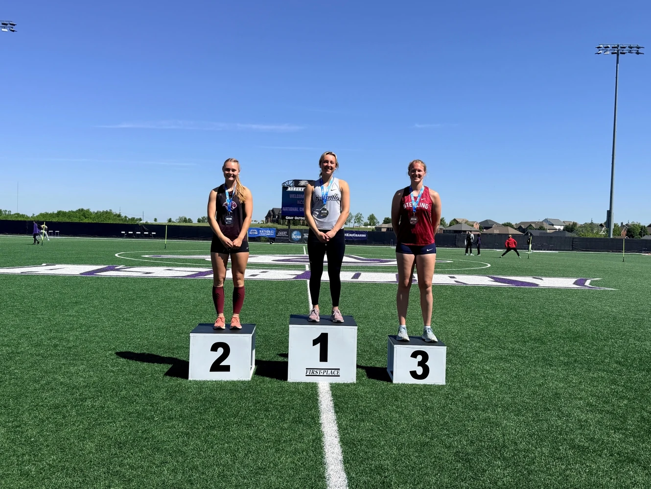 This image depicts three girls standing on podiums ranked from 3rd place to 1st. They are wearing track uniforms. 