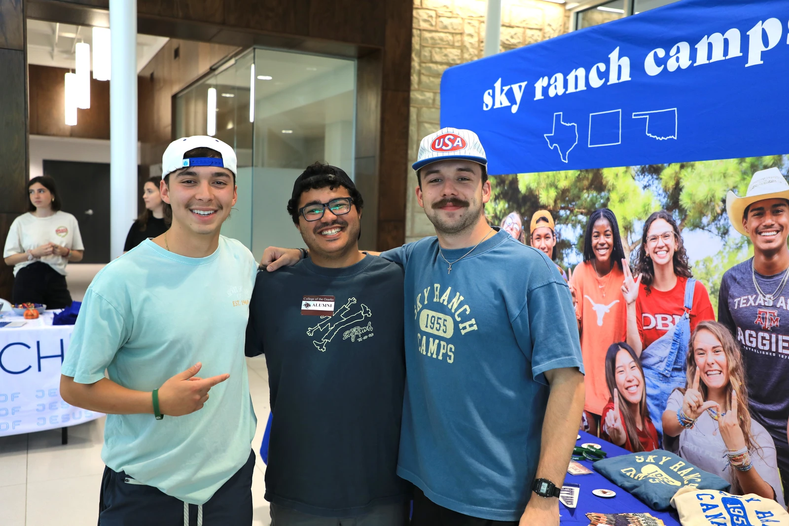 This image depicts three men with their arms around each other smiling at the camera. They are in front of a sign the says, "Sky Ranch Camps". 