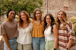 This image depicts five college aged girls standing outside with their arms around each other smiling at the camera. 