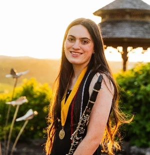 The picture depicts a girl standing outside with the sun glowing behind her. She is wearing a black dress and has graduation cords and a medal around her neck. She has a clarinet in her arm. 