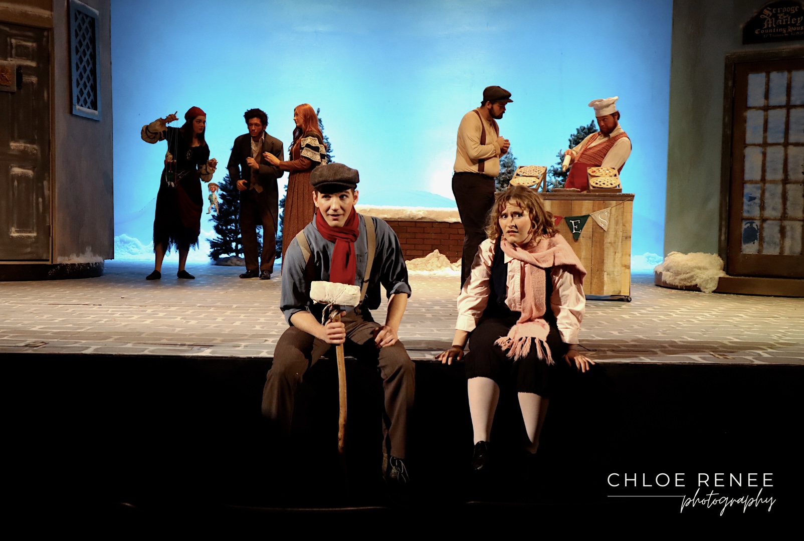 Actors perform on a winter-themed stage set, with two seated at the front and a market stall, snow-covered scenery, and additional performers in the background.