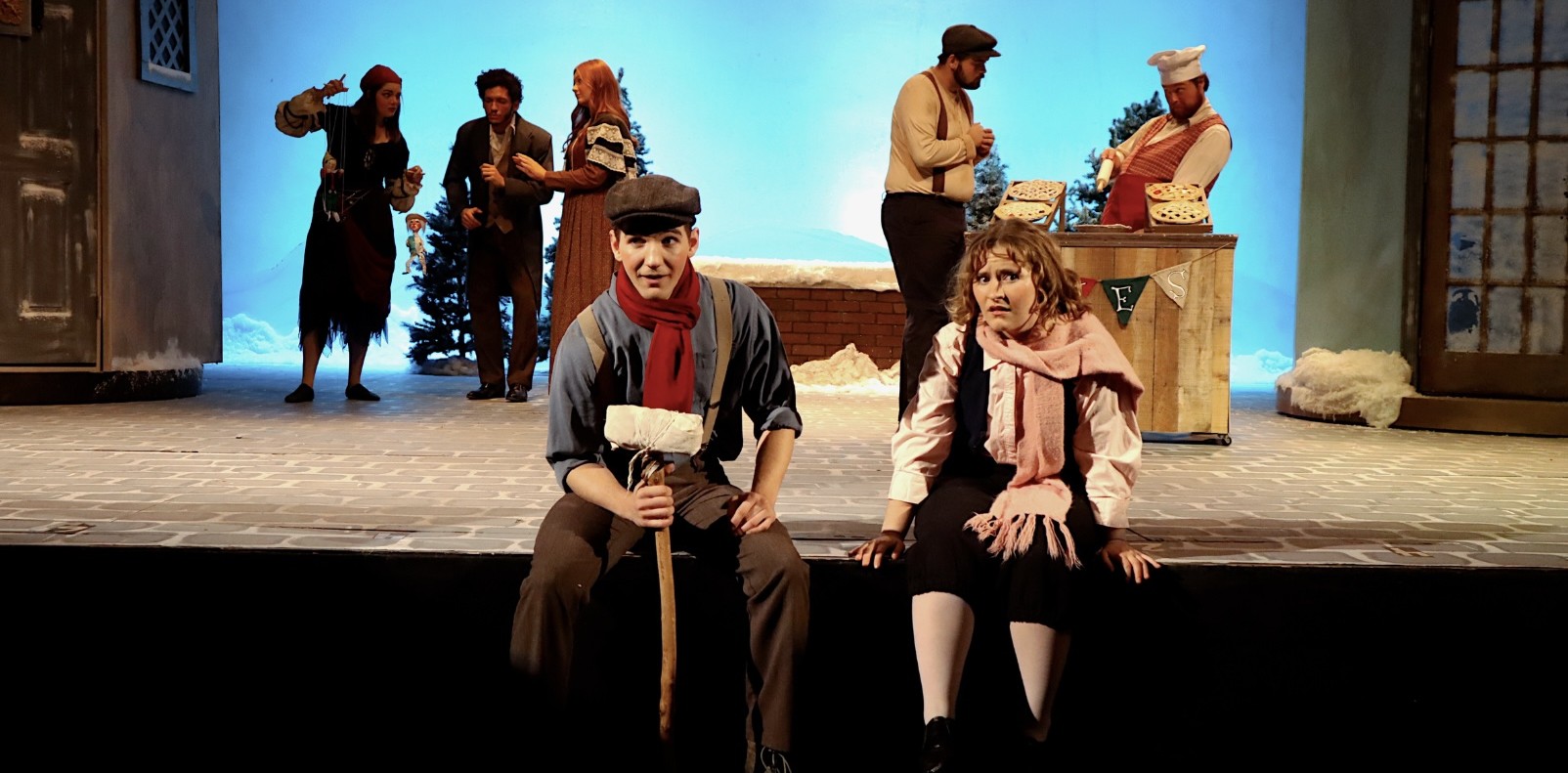 Actors perform on a winter-themed stage set, with two seated at the front and a market stall, snow-covered scenery, and additional performers in the background.
